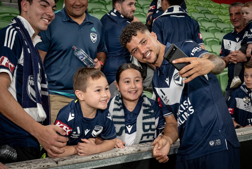 A Melbourne Victory player takes a selfie with two young soccer fans at a match. 