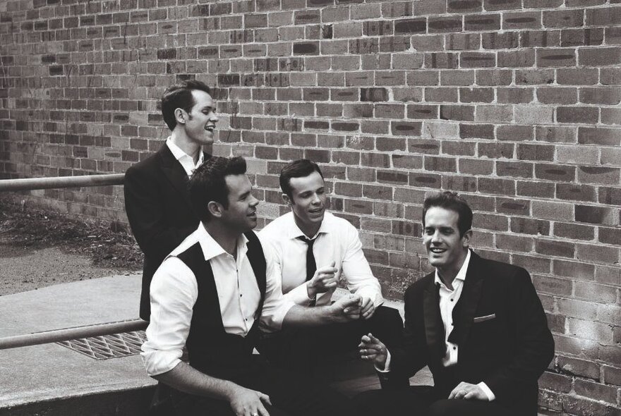 Male four-piece musical group seated on outdoor steps wearing black jackets and white shirts.