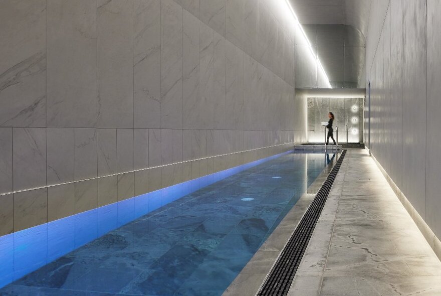 A very long luxury pool in a hotel with lots of marble and blue lights illuminating the water. 