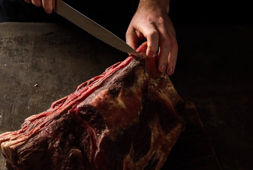 A chef's hand holding a piece of cured meat, with a sharp knife makes a cut.