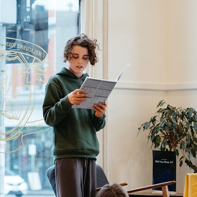 A child holding up a book up and reading in front of a shop front window with traffic lights visible outside. 