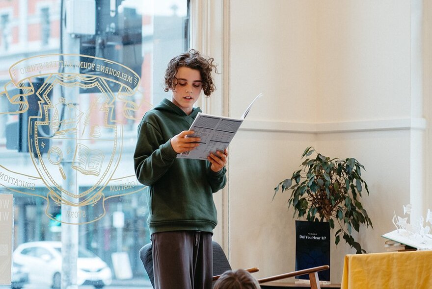 A child holding up a book up and reading in front of a shop front window with traffic lights visible outside. 