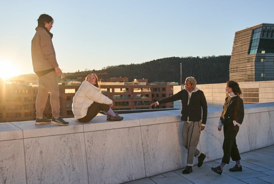 Two people standing on concrete wall, two others looking on, against city backdrop.