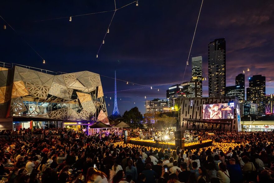 A large crowd of people watching a performance on the stage at Fed Square at night with Melbourne cityscape in the background; nightime.