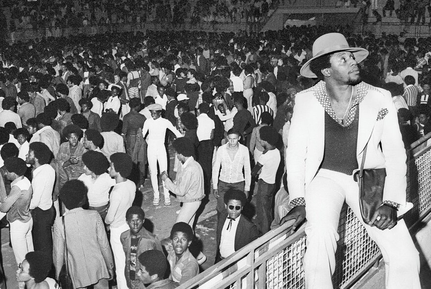 A black and white image of a man in a white suit and large hat sitting on a fence at a concert while people dance and mingle in the background. 