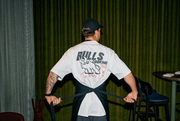 A man wearing a white t-shirt seen from behind, tying apron strings behind his back.