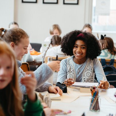 Kids seated around tables writing into notebooks and interacting, during a creative writing workshop in a bright studio space.