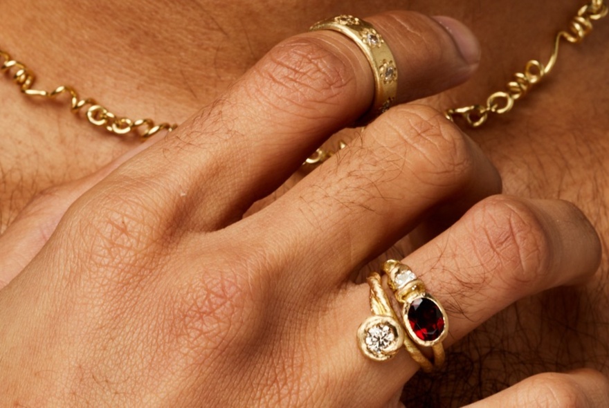 A hand with three gold rings clasping at a gold chain around someone's neck. 