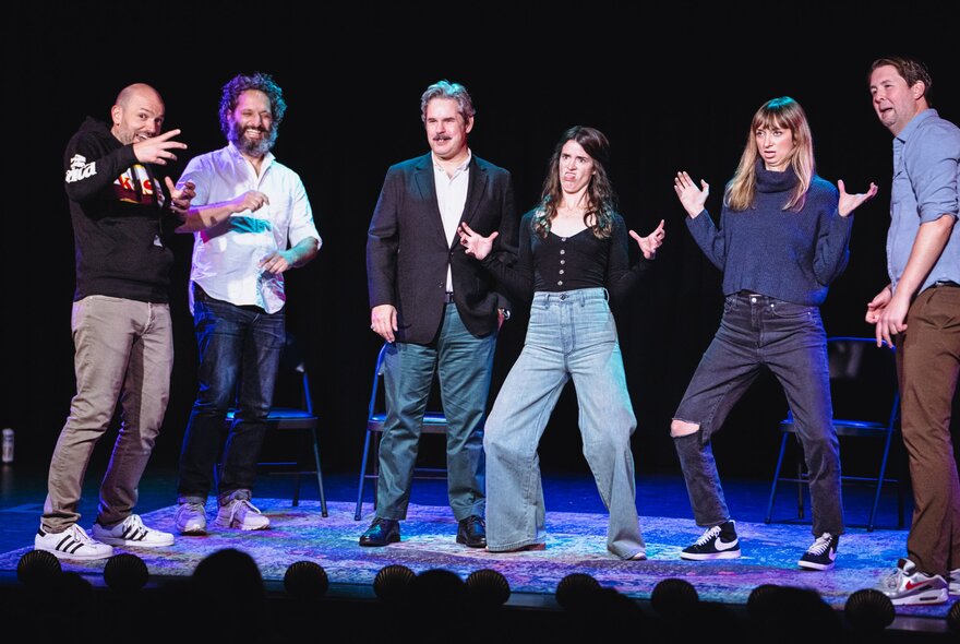 Six performers on stage in front of an audience, improvising a scene and pulling poses and funny faces.