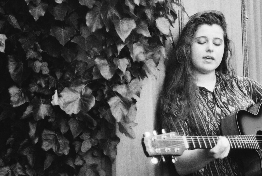 Singer Bridget Allan playing guitar and performing next to some foliage. 
