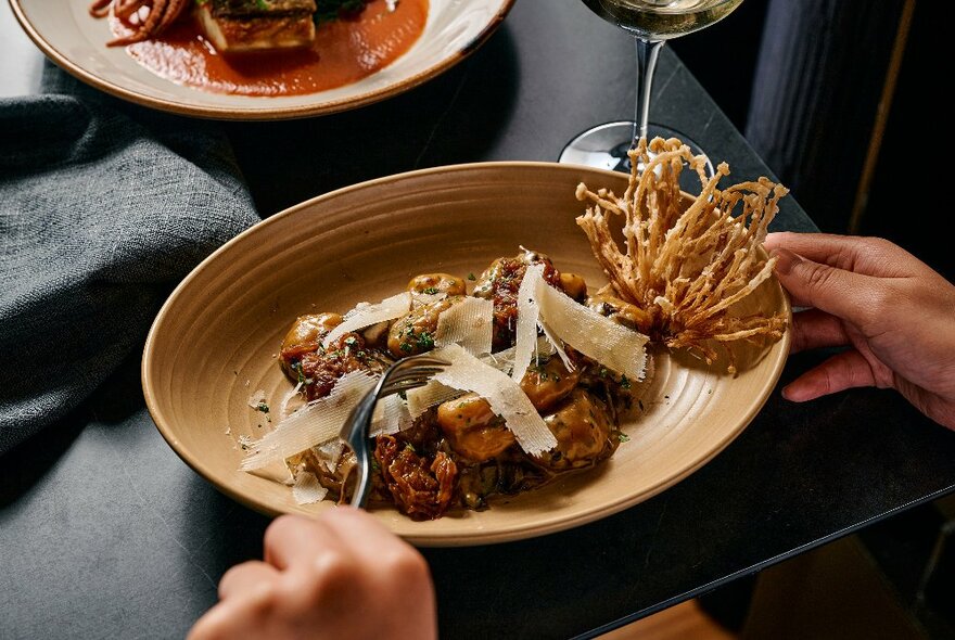 Hands holding a dish of food topped with sliced cheese at a restaurant table.