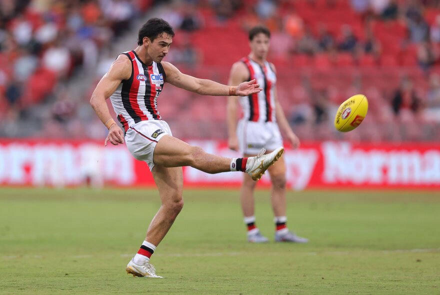 A St Kilda AFL football player on the field with his leg raised high in the air, having just kicked the football, a blurred stadium crowd in the background.