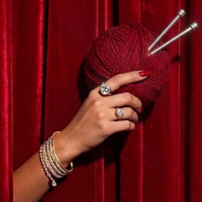 A woman's hand reaching out through red velvet curtains and holding a ball of red wool with two silver knitting needles stuck in it.