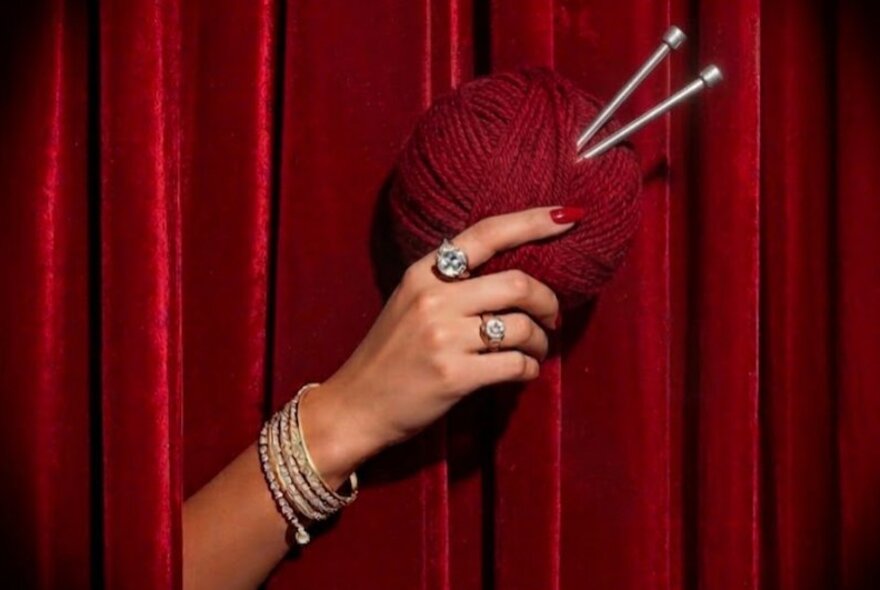 A woman's hand reaching out through red velvet curtains and holding a ball of red wool with two silver knitting needles stuck in it.