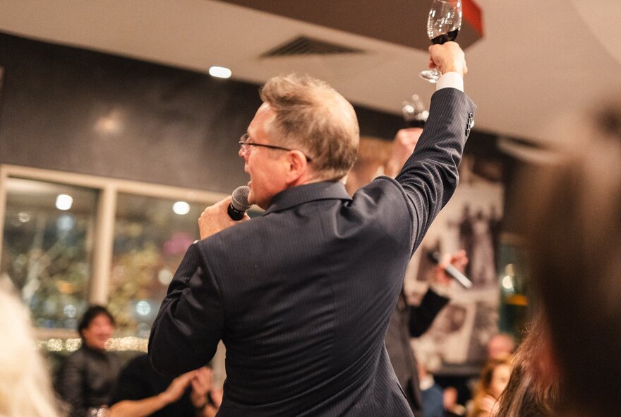 The back of a man with a microphone and one arm raising a glass in a restaurant. 