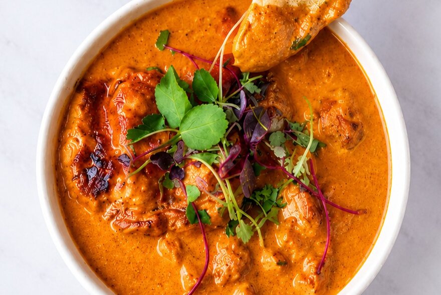 A bowl of creamy tomato-based Indian curry garnished with fresh herbs and microgreens, served alongside a piece of flatbread.