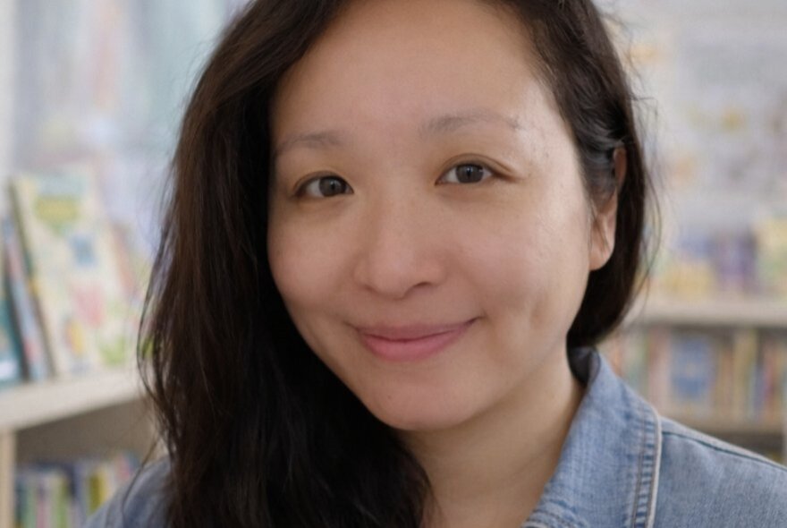 Author Angie Cui, with long dark hair, shelves of children's books in the background.