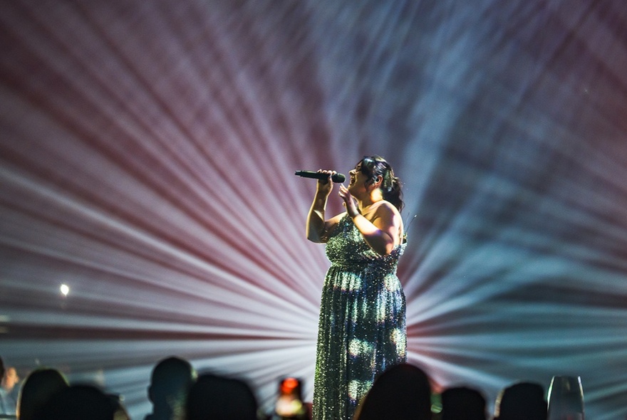 A woman singing into a microphone on the stage at Festival Hall spot lit from behind.