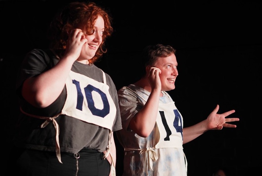 Two individuals wearing bibs with numbers on them, performing impro on a stage, mimicking talking into a phone.