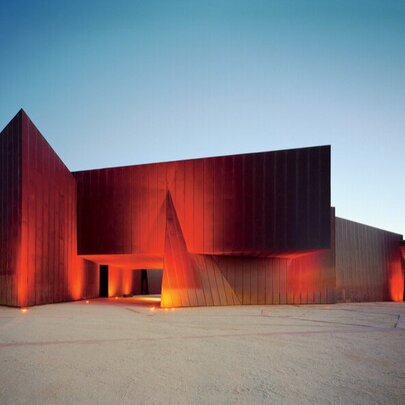 Innovative red-coloured building that is the ACCA art gallery, set against a blue sky, with a large open area in the foreground.