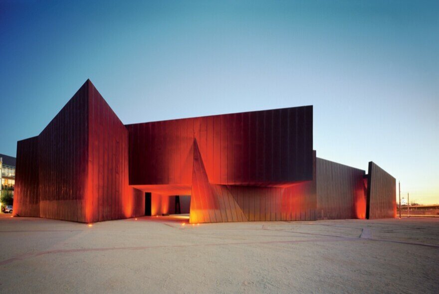 Innovative red-coloured building that is the ACCA art gallery, set against a blue sky, with a large open area in the foreground.