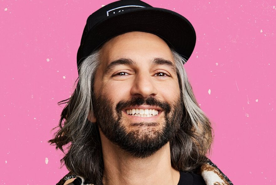 Comedian Amir K, a man with black and silver grey shoulder length hair and a dark beard, smiling at the viewer and wearing a baseball cap, posed against a pink background.