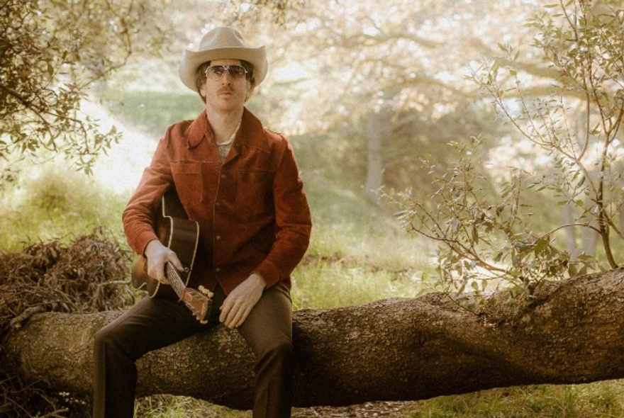 Comedian dressed as a country singer sitting on the branch of a tree holding a guitar, wearing sunglasses, cowboy hat and brown jacket in leafy garden.