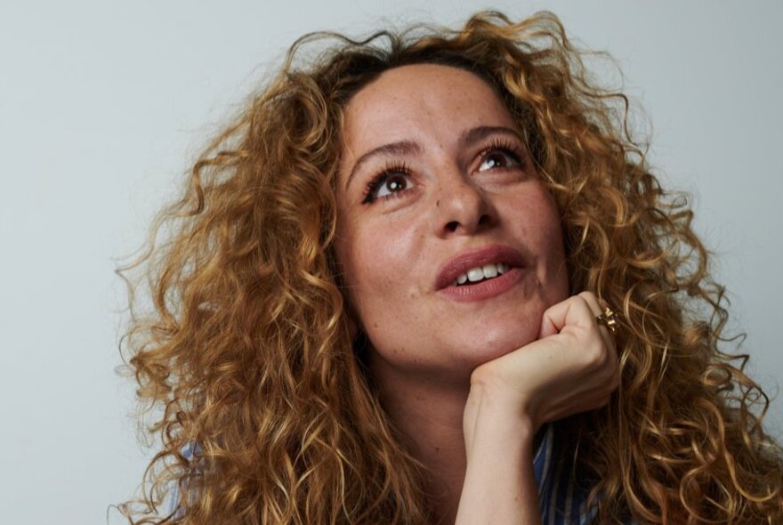 A woman with long, wild, curly hair leaning her chin on her hand while looking upwards and smiling.