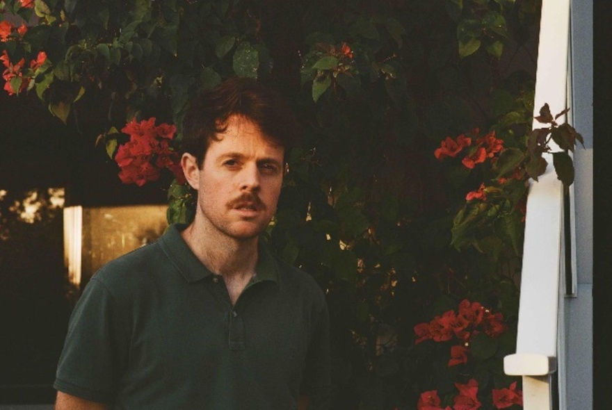 Young man with moustache and dark hair, wearing a dark green polo shirt standing in a garden with dark red floral plants.