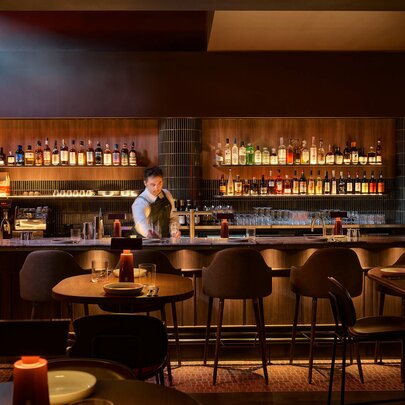 Interior of a bar, with a bartender behind a timber counter, a wall of bottled drinks behind him, high bar stools and some tables in the foreground.
