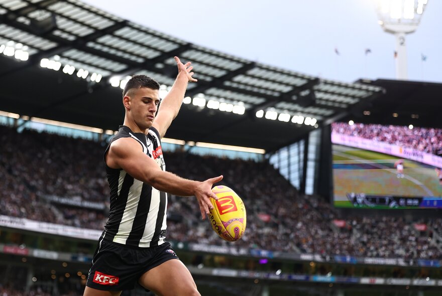 A Collingwood AFL player running while about to kick a football, one arm stretched high above his head, with a large stadium crowd in the background. 