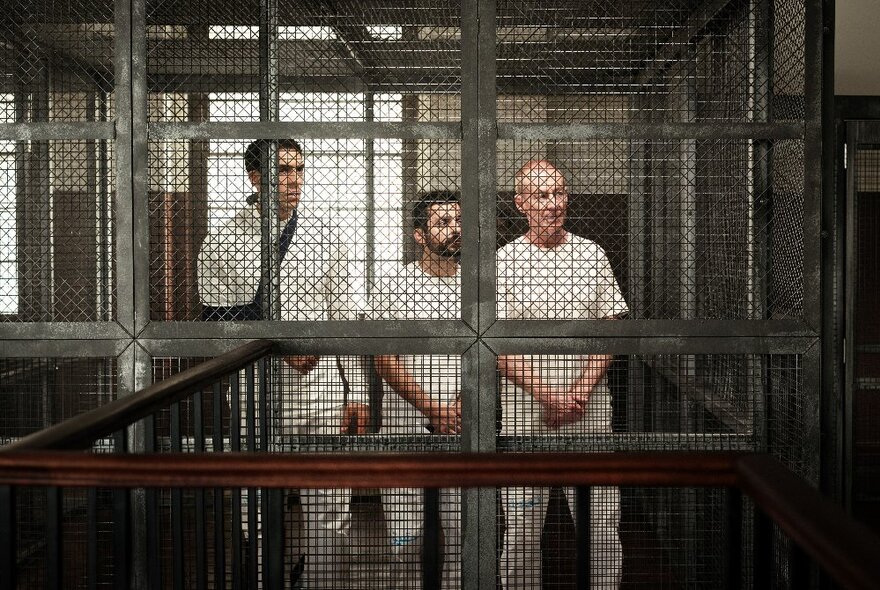 A still from the movie The Correspondent showing three men in a wire cage in prison. 