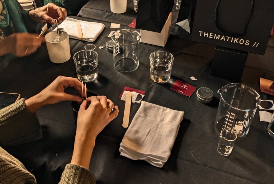 Hands holding a wick over a candle pot with glasses of water and workshop implements.