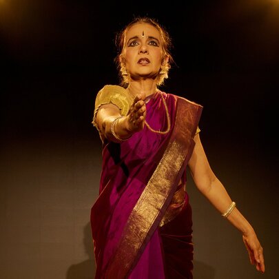 An Indian woman wearing a magenta sari with gold trim, traditional jewellery and face adornments, on stage.