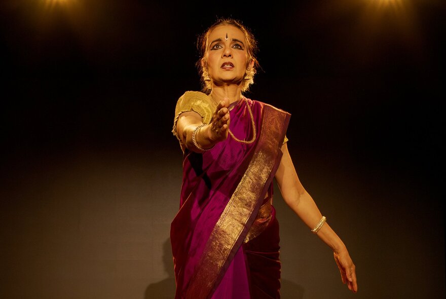 An Indian woman wearing a magenta sari with gold trim, traditional jewellery and face adornments, on stage.
