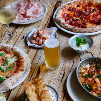 A wooden table in a restaurant with plates of pizza, salad, bread, cured meats and drinks.