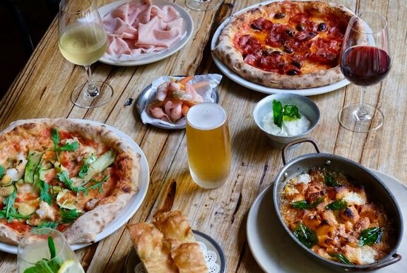 A wooden table in a restaurant with plates of pizza, salad, bread, cured meats and drinks.