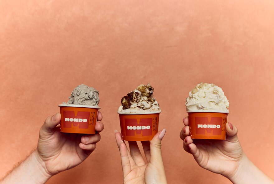 Three hands hold small orange cups filled with different flavours of ice cream against a pale orange background.