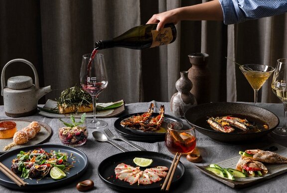 A person pouring red wine into a wine glass on a table with a variety of dishes including seafood, salads and desserts.