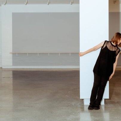 A mature-age dancer in black, leaning sideways while holding onto a white column in a dance studio.