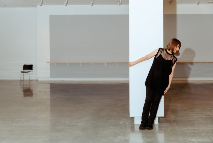 A mature-age dancer in black, leaning sideways while holding onto a white column in a dance studio.