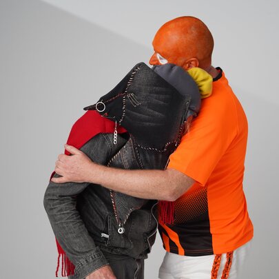 Video still from a film, two people hugging against a plain, light grey background, with both their faces covered.