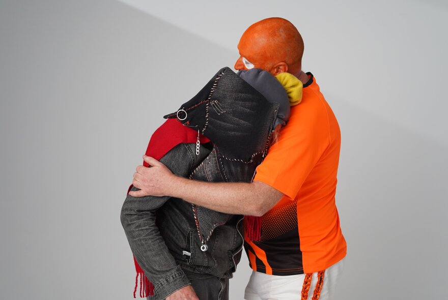 Video still from a film, two people hugging against a plain, light grey background, with both their faces covered.