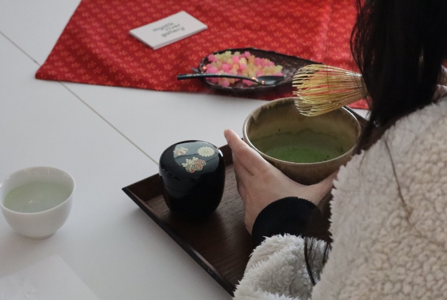 Looking over the shoulder of an Asian woman at a bowl of traditional Japanese tea, as she prepares to whisk it with a traditional Japanese Tea whisk.