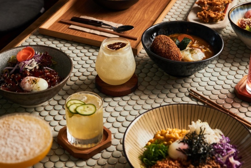 A table at a restaurant with bowls of Japanese food, drinks, cutlery and chopsticks.