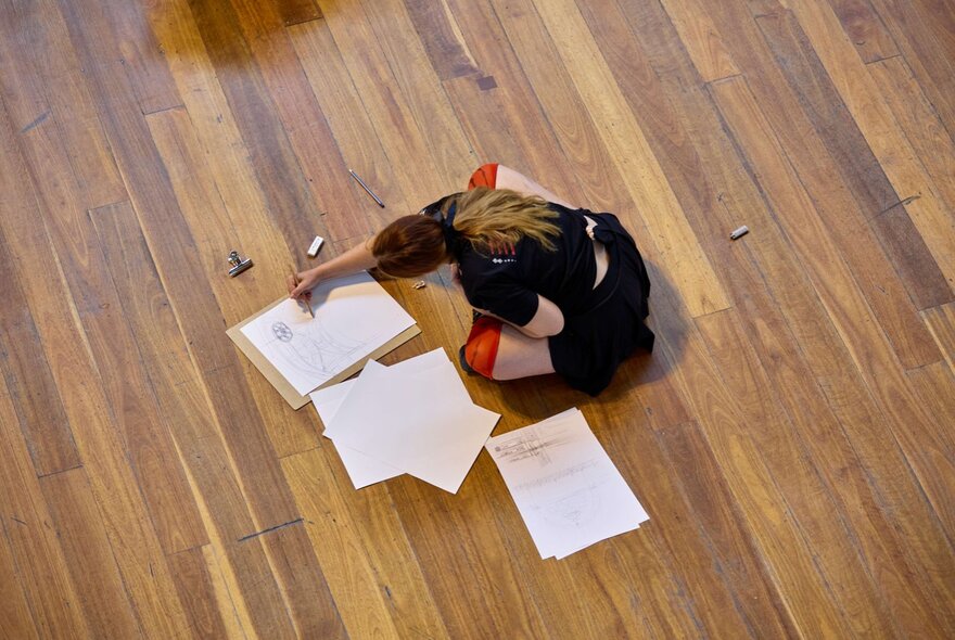 A woman sitting cross-legged on the wooden floor of the Royal Exhibition Building, Melbourne, drawing.