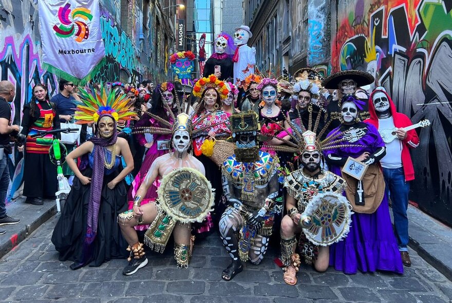 A group of people posed in a city laneway, dressed in Day of the Dead costumes with matching day of the Dead painted faces, some holding instruments.