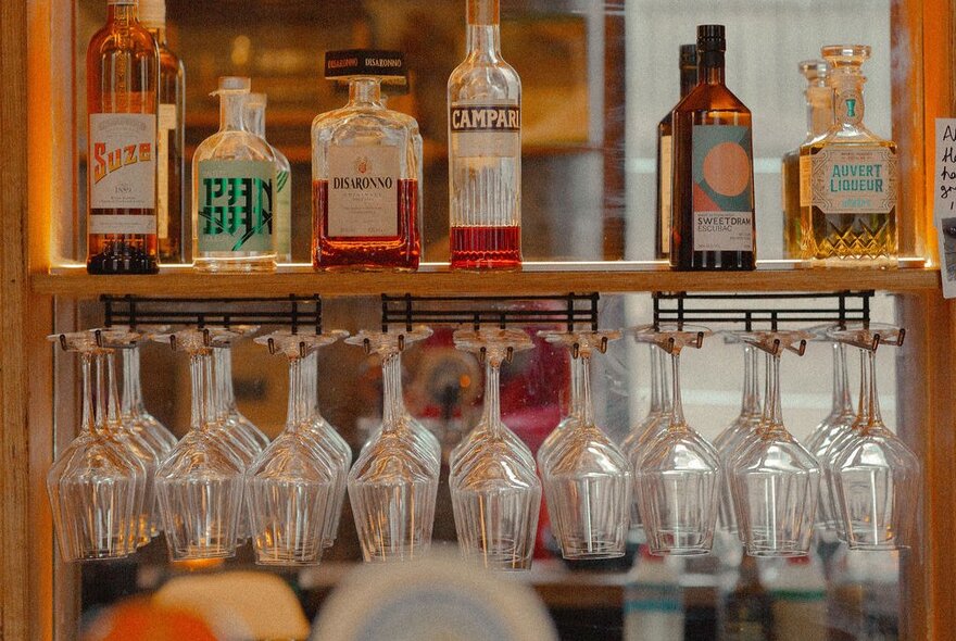 A bottle lineup one bar shelf with glasses hanging upside down. 