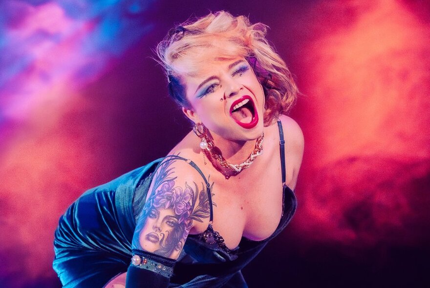 A burlesque dancer with tattoos performing, bent over and her mouth wide open with an orange, fiery glow in the background. 