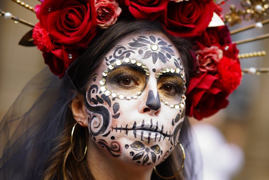 A person with an intricate and elaborate painted Day of the Dead face, and a red rose headdress with a black veil.